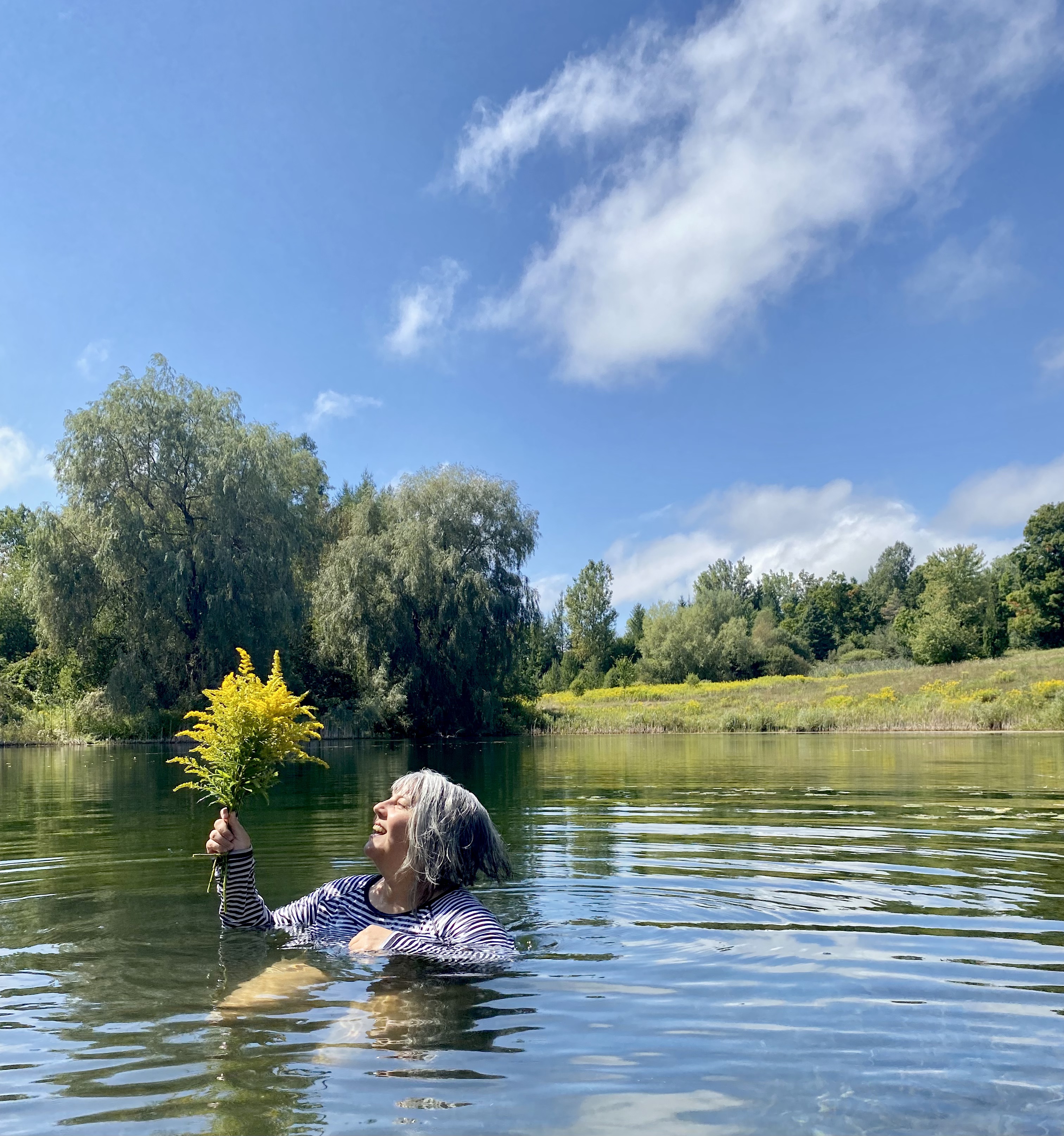 Anna floating in a pond wearing a striped black and white shirt, holding a bunch of yellow flowers and looking at them. Trees and green grass in the background.